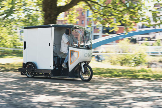 ONO Cargobike fährt bei schönem Wetter auf Fußgängerweg an einem Fluß entlang
